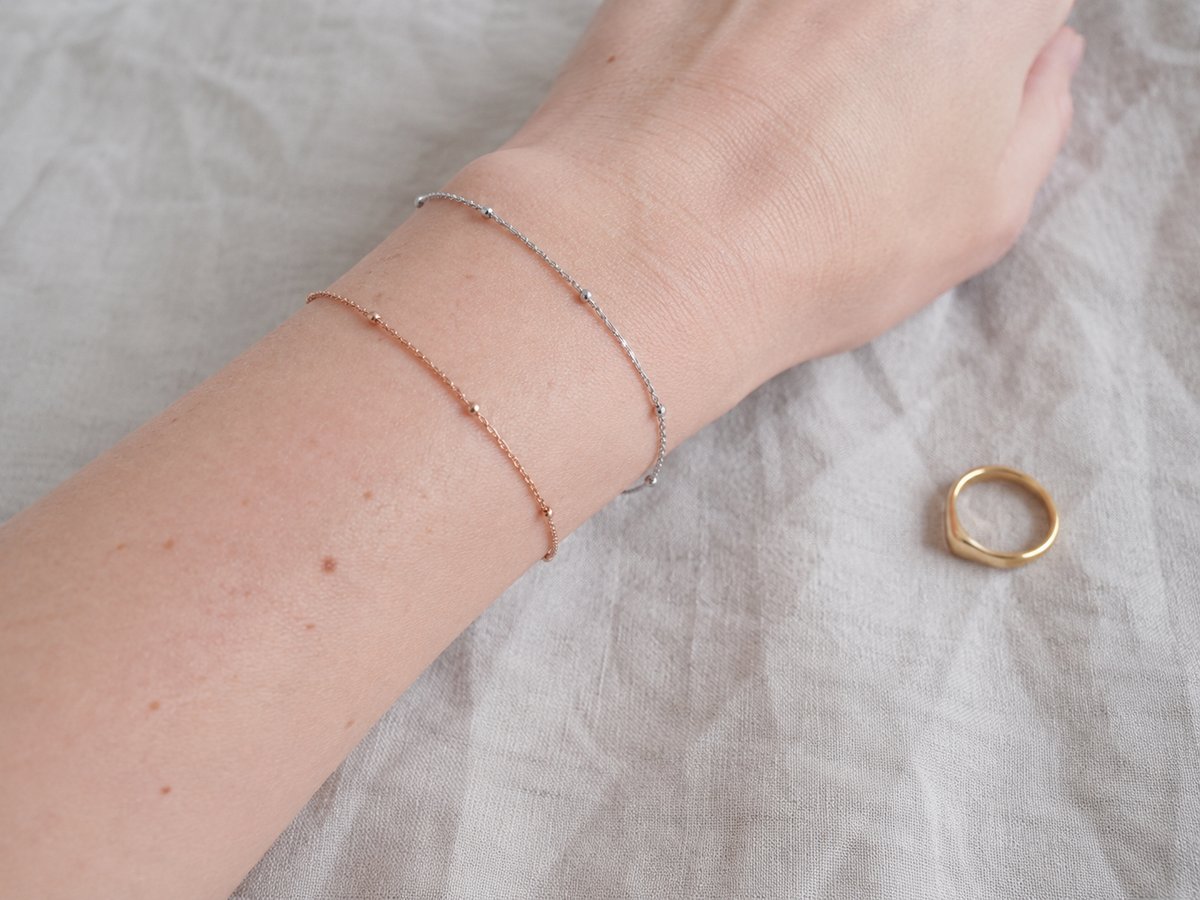 Handgelenk mit zwei zarten Armbändern in Silber und Roségold, mit einem kleinen goldenen Ring auf einem beigen Stoff