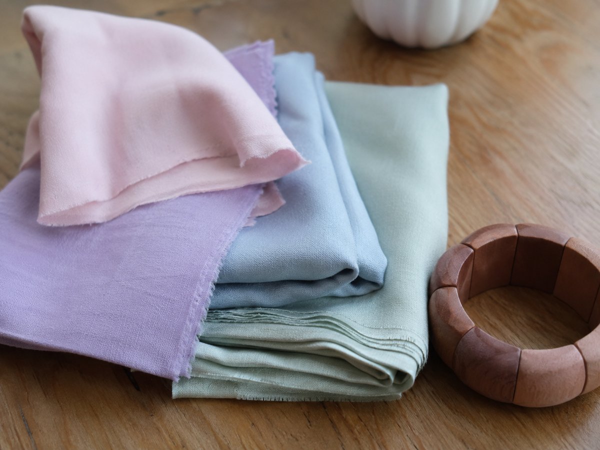Gefaltete Stoffmuster in Pastelltönen Rosa, Lila, Blau und Salbeigrün auf einer Holzoberfläche mit einem Holzarmreif